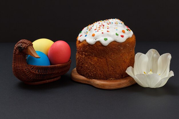 Gâteau de Pâques sucré sur une planche à découper en bois et des œufs de Pâques colorés dans un panier en osier avec une fleur de tulipe sur la surface noire