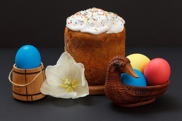 Gâteau de Pâques sucré sur une planche à découper en bois et des œufs de Pâques colorés dans un panier en osier avec la surface noire