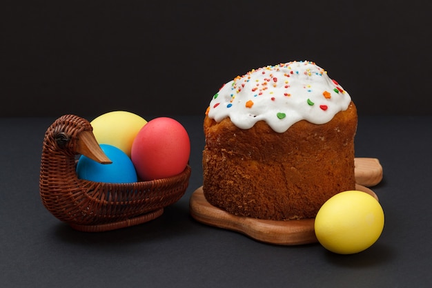 Gâteau de Pâques sucré sur une planche à découper en bois et des œufs de Pâques colorés dans un panier en osier avec la surface noire