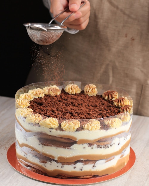 Gâteau de type tiramisu à la main féminine avec de la poudre de cacao. Nature morte avec l'ensemble du gâteau et des grains de café sur une table en bois.