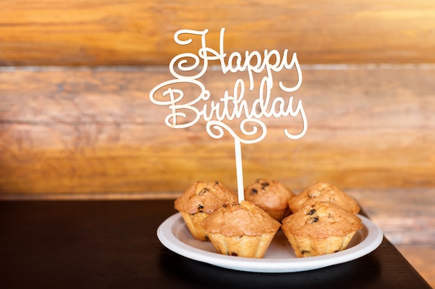 Photo gâteaux d'anniversaire et muffins avec panneau de voeux en bois sur mur rustique. chante en bois avec des lettres joyeux anniversaire et bonbons de vacances.