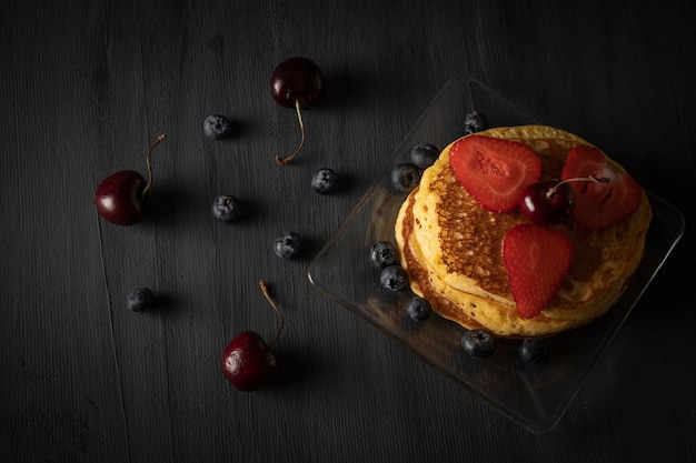 Gâteaux chauds empilés avec des fraises de cerises et des myrtilles sur une table en bois noire de cru