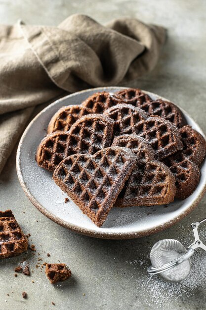 Gaufres au chocolat en forme de coeur saupoudré de sucre en poudre dans une assiette en céramique sur fond texturé avec une serviette en lin au premier plan, nature morte pour la Saint-Valentin