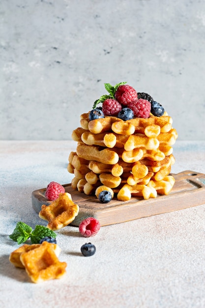 Gaufres belges croustillantes fraîches pour le petit déjeuner avec des baies mûres (framboises, myrtilles, mûres), menthe et sucre en poudre sur une planche de bois sur un fond clair.