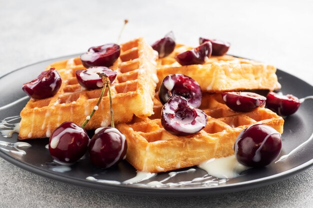 Gaufres cannelées belges avec cerises fraîches et crème sur une assiette. Gaufres maison douces, dessert sucré aux cerises.