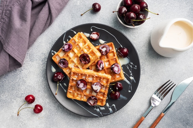 Gaufres cannelées belges avec cerises fraîches et crème sur une assiette. Gaufres maison douces, dessert sucré aux cerises.