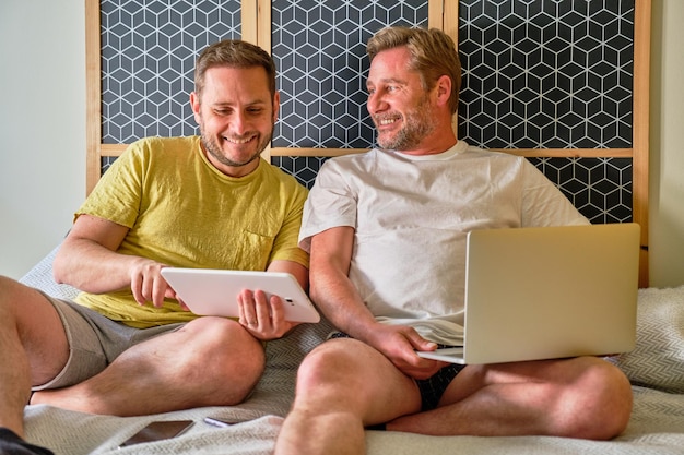 Gay mature couple sitting on bed smiling regardant quelque chose sur tablette et ordinateur portable