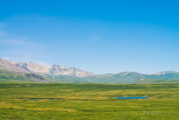 Géant des montagnes de neige au-dessus d'une vallée verdoyante sous un ciel bleu clair. Prairie avec une riche végétation et des lacs de hautes terres au soleil. Magnifique paysage de montagne ensoleillé de nature majestueuse.