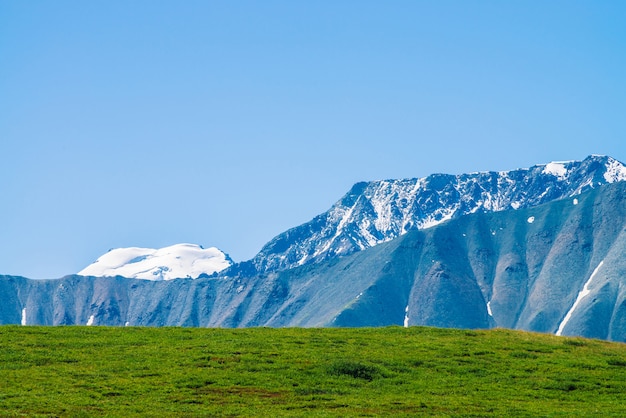 Géant des montagnes avec de la neige au-dessus de la vallée verte en journée ensoleillée. Glacier sous le ciel bleu Prairie avec une riche végétation de hautes terres au soleil. Magnifique paysage de montagne enneigée d'une nature majestueuse.