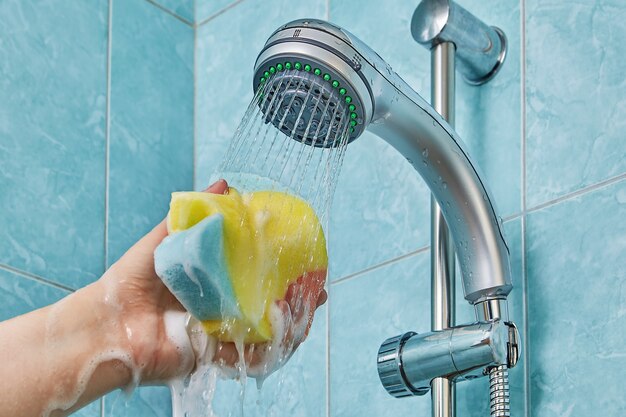 Le gel douche mousse et coule à travers l'éponge pour la salle de bain qui est comprimée dans la main humaine étirée sous le flux d'eau s'écoulant de la pomme de douche.