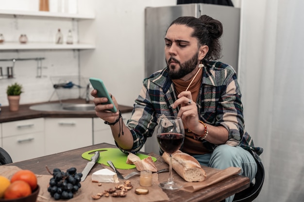 Génération moderne. Gentil homme réfléchi naviguant sur Internet sur son smartphone tout en mangeant du fromage