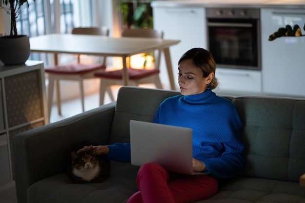 Genre femme caucasienne caressant un chat distrait du travail indépendant avec un ordinateur portable sur les genoux est assis sur un canapé