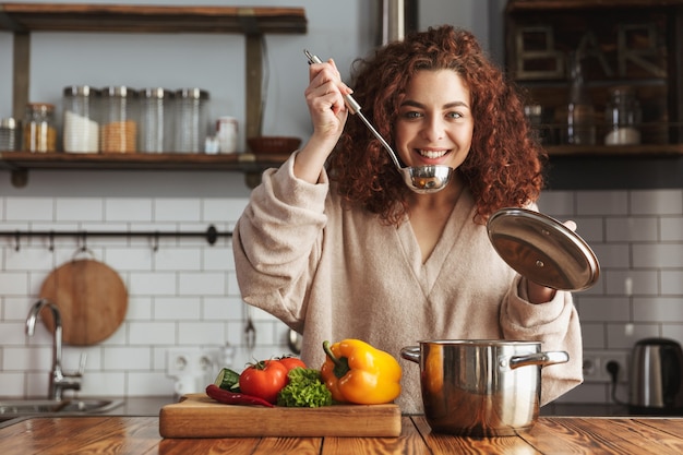 genre femme caucasienne tenant une cuillère de louche de cuisson tout en mangeant de la soupe avec des légumes frais dans la cuisine à la maison