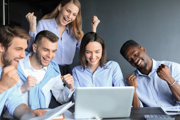 Des gens d'affaires heureux rient près d'un ordinateur portable au bureau. Les collègues de l'équipe qui réussissent plaisantent et s'amusent ensemble au travail.