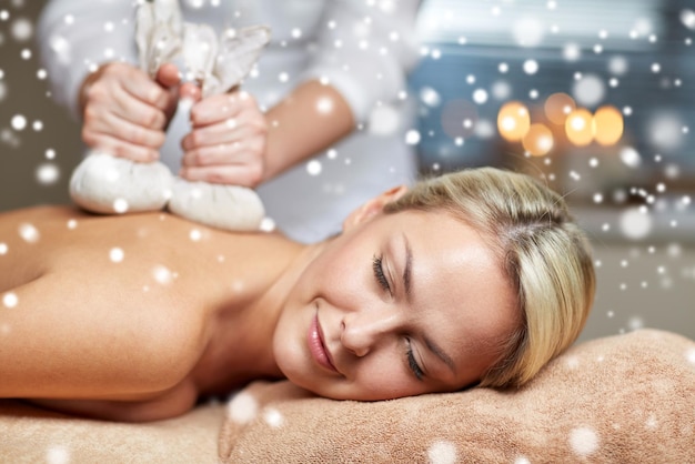 gens, beauté, spa, hiver et concept de détente - gros plan d'une belle jeune femme allongée et ayant un massage du dos avec un sac à base de plantes dans un salon de spa avec effet de neige