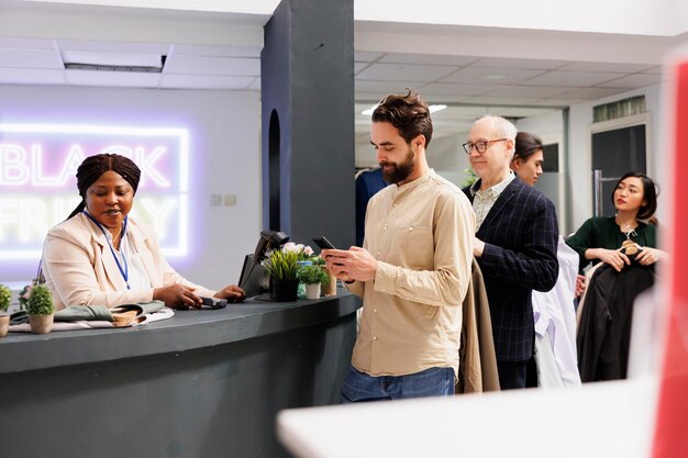 Des gens divers se tiennent en ligne avec des vêtements à la main tout en faisant leurs courses le vendredi noir dans un magasin de mode. Les clients attendent le paiement à la caisse enregistreuse, achetant des vêtements à des prix réduits