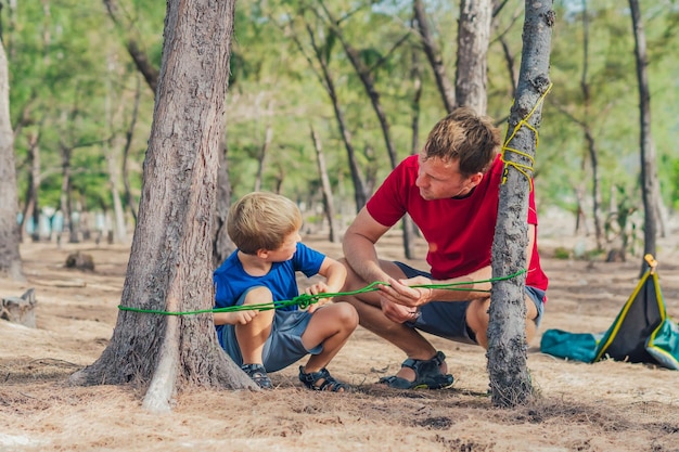 Les gens du camping les touristes de style de vie en plein air dans la forêt d'été près de la mer de lazur Garçon blond fils avec le père étudient les techniques de survie pratiquent les méthodes de faire des nœuds de corde Éducation naturelle des enfants