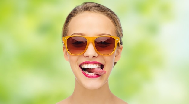 gens, expression, joie et concept de mode - jeune femme souriante à lunettes de soleil avec rouge à lèvres rose sur les lèvres montrant la langue sur fond vert d'été