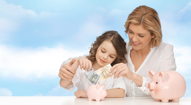 les gens, les finances, le budget familial et le concept d'épargne - heureuse mère et fille avec des tirelires et du papier-monnaie sur fond de ciel bleu