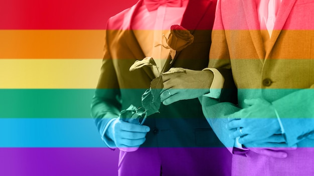 les gens, l'homosexualité, le mariage homosexuel et le concept d'amour - gros plan d'un couple gay heureux avec une fleur de rose rouge tenant la main sur le mariage sur fond de rayures de drapeau arc-en-ciel