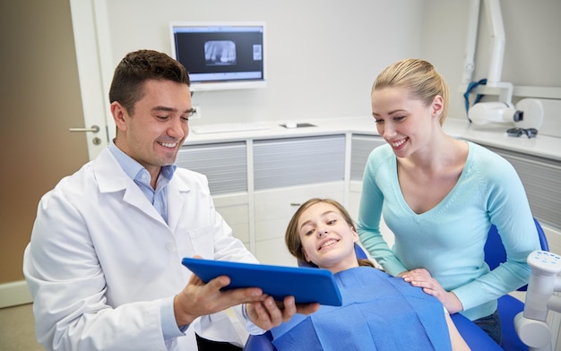 les gens, la médecine, la stomatologie, la technologie et le concept de soins de santé - un dentiste masculin heureux montrant un ordinateur tablette à une patiente et à sa mère au bureau de la clinique dentaire