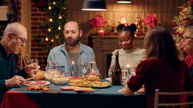 Des gens multiculturels joyeux appréciant le dîner de Noël tout en mangeant du poulet rôti. Heureux membres de la famille diversifiée appréciant la cuisine traditionnelle faite maison tout en célébrant ensemble les vacances d'hiver.