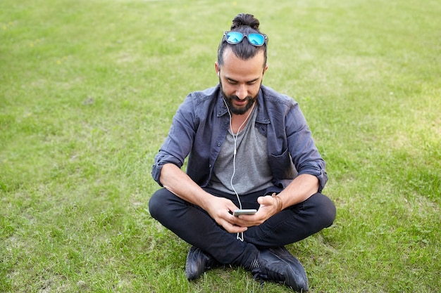 gens, musique, technologie, loisirs et style de vie - jeune homme hipster heureux avec écouteurs et smartphone assis sur l'herbe