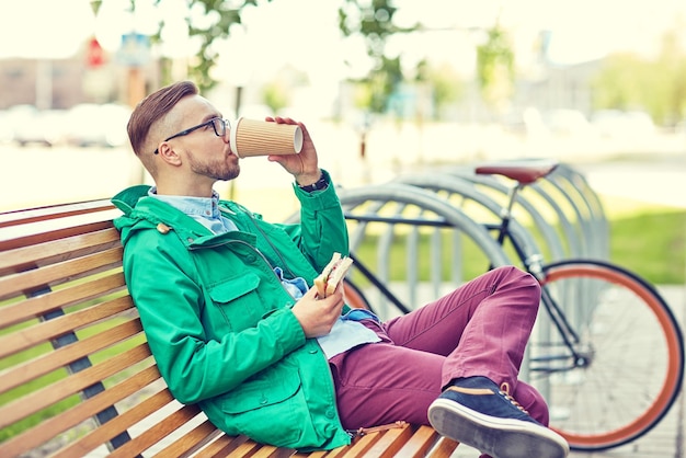 les gens, le style, les loisirs et le mode de vie - un jeune hipster heureux buvant une tasse de café et mangeant un sandwich dans la rue de la ville