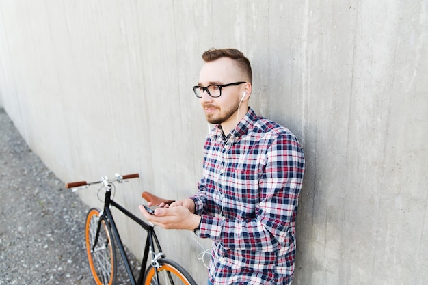 les gens, le style, la technologie, les loisirs et le style de vie - jeune homme hipster heureux dans les écouteurs avec smartphone et vélo à pignon fixe écoutant de la musique dans la rue de la ville