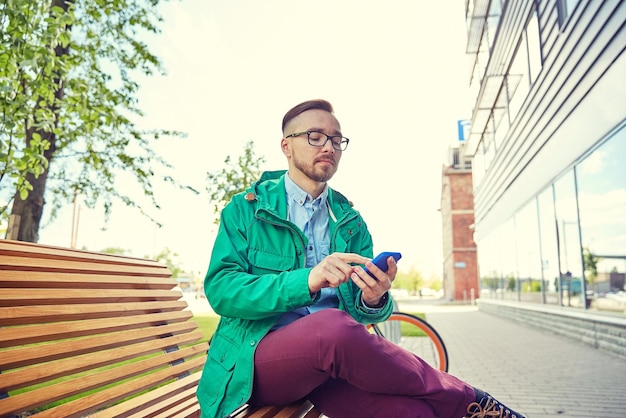 les gens, le style, la technologie et le style de vie - jeune homme hipster heureux avec smartphone et vélo fixie assis sur un banc en ville