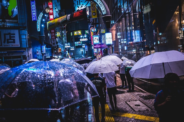 Des gens tenant des parapluies en marchant dans la rue au milieu des bâtiments la nuit.