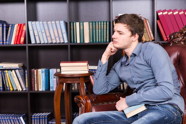 Gentil beau jeune homme assis sur la chaise de la bibliothèque tout en s'appuyant sur son bras et en regardant vers la gauche du cadre.