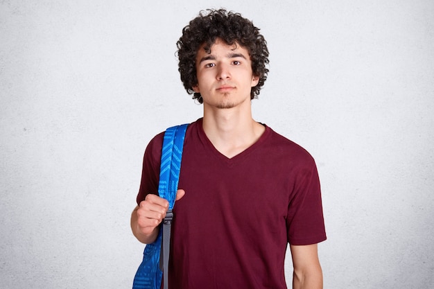 Gentil beau mec aux cheveux noirs bouclés, debout, ayant un sac à dos bleu sur le dos, portant un t-shirt rouge foncé