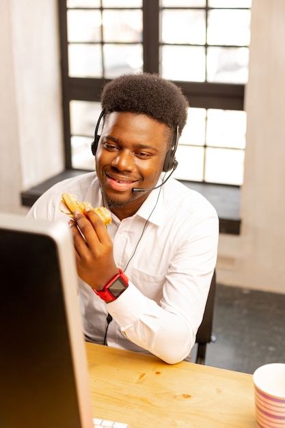 Gentil homme positif assis devant l'ordinateur avec un sandwich en mangeant au travail