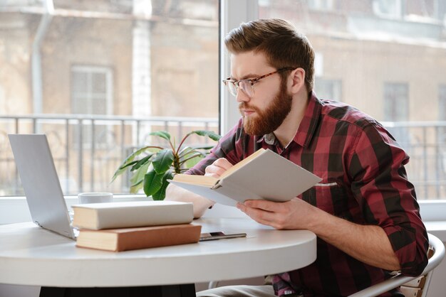 Gentil jeune homme barbu lisant des livres tout en utilisant un ordinateur portable