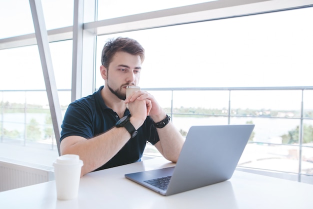 Gentil jeune homme est assis à la table avec un smartphone dans ses mains et regarde l'écran du portable