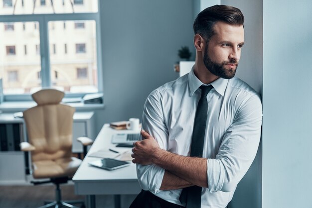 Gentil jeune homme en tenue de soirée en détournant les yeux et en gardant les bras croisés en se tenant debout au bureau