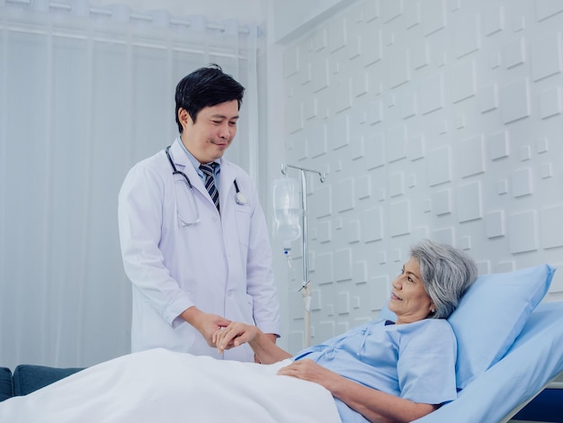 Le gentil médecin asiatique en costume blanc visite des entretiens et apporte son soutien en tenant la main d'une patiente âgée heureuse en robe bleu clair allongée sur un lit dans une solution saline dans une chambre d'hôpital