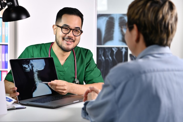 Gentil réconfortant et attentionné. Souriant jeune médecin asiatique en uniforme chirurgical consultant et expliquant une patiente à l'aide d'un ordinateur portable dans la salle d'examen.