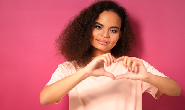 Photo geste d'amour avec un regard tendre belle jeune femme regardant positivement à l'avant portant un t-shirt pêche isolé sur un mur rose