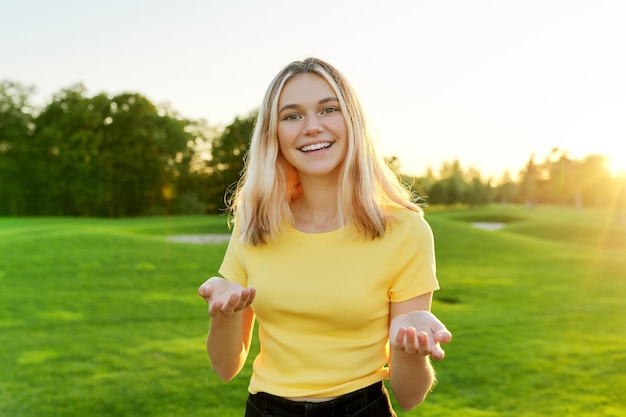 Gestes de jeune belle adolescente parlant à la caméra, étudiante de 16, 17 ans donnant une interview en regardant la caméra, pelouse verte au coucher du soleil sur fond de parc