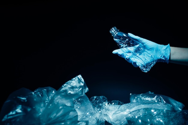 La gestion des déchets. Recyclage du plastique. Problème d'écologie. Protection environnementale. Main bénévole dans un gant bleu collectant une bouteille usagée à partir d'une pile de sacs en cellophane isolée sur fond d'espace de copie sombre.