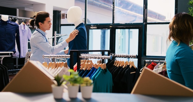 Gestionnaire asiatique fermant les boutons de costume bleu, organisant des vêtements à la mode et des accessoires élégants dans une boutique moderne. Joyeux employé travaillant dans un magasin de vêtements visuel. Notion de mode