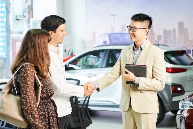 Gestionnaire asiatique à lunettes souriant et saluant jeune couple avec poignée de main dans la salle d'exposition de voiture