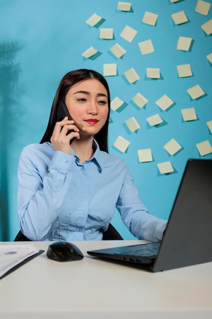 Gestionnaire asiatique parlant sur un téléphone intelligent pendant la journée de travail, utilisant la communication à distance pour parler avec des collègues. Femme professionnelle dans un ordinateur portable de bureau contemporain.