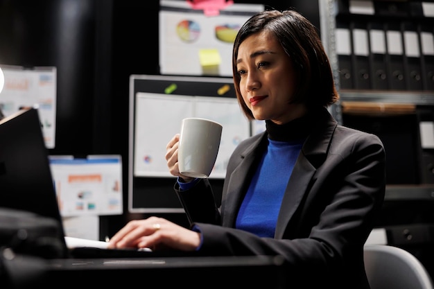 Gestionnaire de femme d'affaires de comptabilité joyeuse appréciant une tasse de café et imputant des données de plan budgétaire sur un ordinateur portable dans un bureau d'affaires. Employé asiatique dans une salle de classeur avec des dossiers sur des étagères