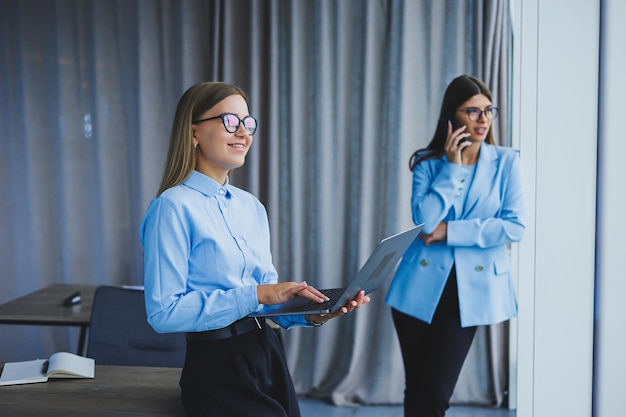 Gestionnaire femme dans des verres classiques souriant pendant le temps de travail au bureau femme européenne positive en chemise bleue bureau avec ordinateur portable travail à distance Un collègue est en arrière-plan mise au point sélective