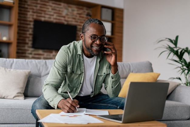 Gestionnaire de gars afro-américain mature souriant occupé dans des verres et décontracté avoir un appel par téléphone fonctionne sur un ordinateur portable