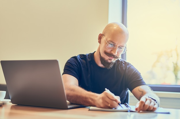 Gestionnaire d'homme chauve adulte concentré utilisant un ordinateur portable tout en écrivant des notes dans un ordinateur portable sur le lieu de travail