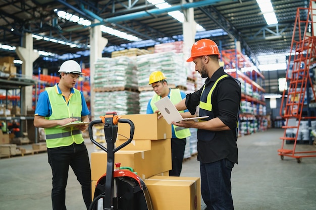 Gestionnaire homme utilisant un ordinateur portable vérifier le stock de carton sur un chariot élévateur et un collègue à l'entrepôt Travailleur portant des vêtements à haute visibilité et un casque de sécurité pour vérifier le comptage des marchandises ou des boîtes pour la livraison